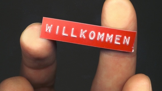 Ein Klebeschild mit der Aufschrift Willkommen wird auf zwei Fingern in die Kamera gehalten. © photocase.de Foto: una.knipsolina
