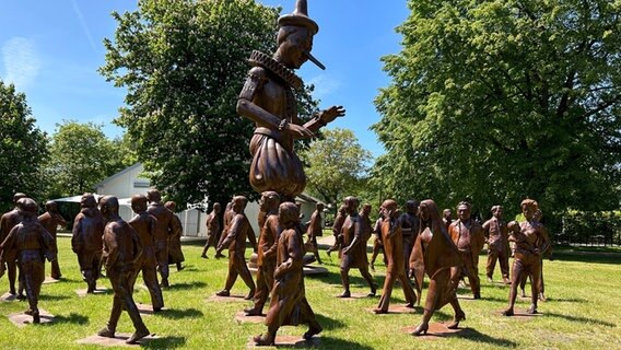 Um eine große hölzerne Pinnochio-Figur sind Menschen-Figuren in einem Kreis aufgestellt. © NDR Foto: Hauke Sievers