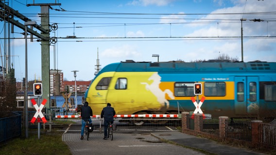 Nahverkehrszug rollt über Bahnschienen. Fahradfahrer warten vor geschlossener Schranke. Der Zug ist in Bewegung, deshalb ist er im Foto leicht unscharf zu erkennen. © Lornz Lorenzen Foto: Lornz Lorenzen