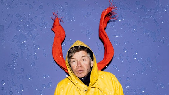 Ein Mann in Regenmantel und mit rotem Schal schaut in die Kamera. Er sieht aus als würde er die Luft anhalten. Um Ihn herum sind Regentropfen. © Sinje Hasheider Foto: Sinje Hasheider