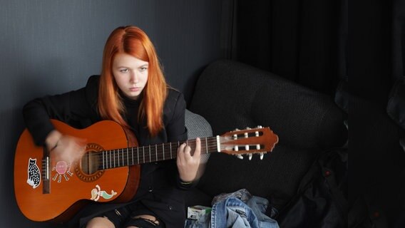 Eine junge Frau mit rotem Haar sitzt vor Schwarzer Wand auf dunklem Sofa und spielt Gitarre. © Julia Solovieva 