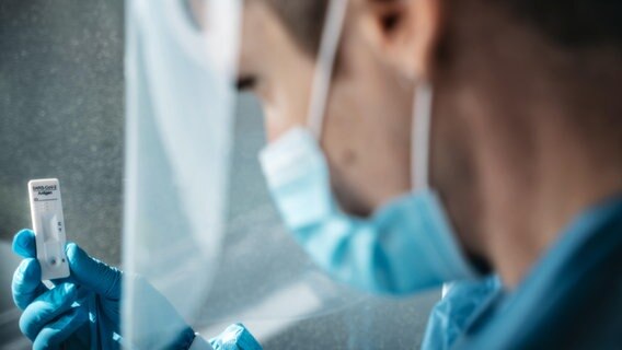 Arzt oder Krankenpfleger hält eine Schnelltest Kasette in der Hand während er Schutzkleidung, Maske und Gesichtsschutz während der Arbeit trägt. © picture alliance / CHROMORANGE Foto: Michael Bihlmayer