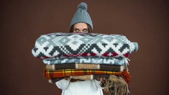 Eine junge Frau mit Pudelmütze hält einen Stapel Wolldecken. © colourbox Foto: -