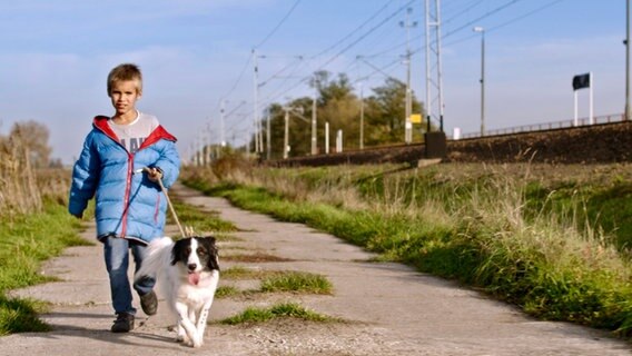 Ein neunjähriger Junge namens Andryi geht mit seinem Hund an der Leine einen Weg entlang. Ländliche Umgebung. Der Hund ist mittelgroß, hat ein schwarz-weißes Fell, seine Name ist Slava. © NDR/Red Balloon 