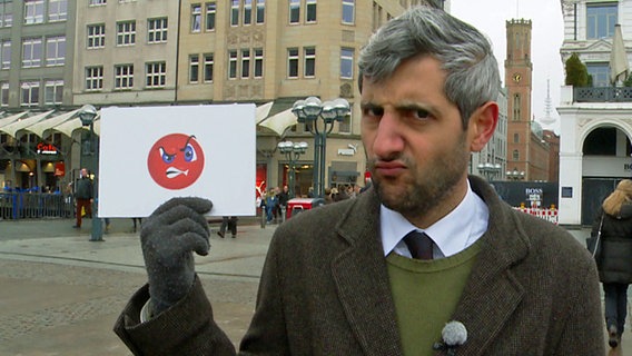Michel Abdollahi guckt zerknirscht und hält ein Schild mit einem wütenden Smiley in der Hand. © NDR / Stefan Mühlenhoff 