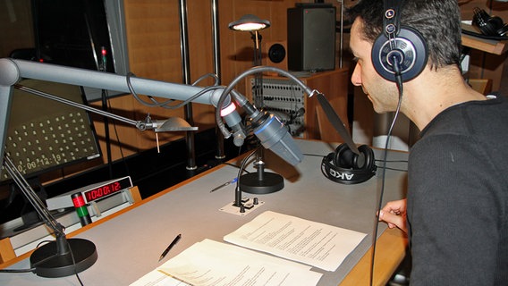 Tarek Youzbachi bei der Sprachaufnahme einer Audiodeskription. Er hat kurze dunkle Haare, trägt Kopfhörer und sitzt an einem Tisch vor einem Mikrofon. © NDR Foto: Martin Ovelgönne