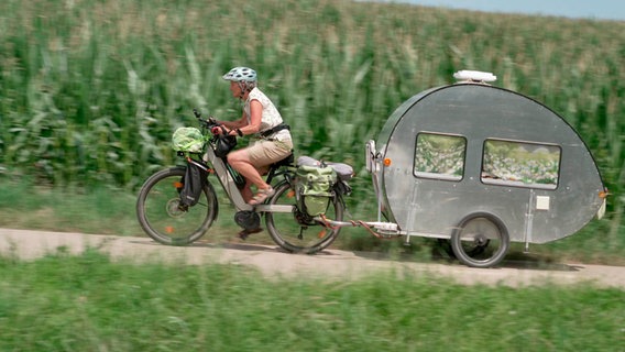 Wohnwagen im Miniformat: Eine Fahrradfahrerin zieht ihr mobiles Zuhause als Anhänger hinter sich her. © WDR/NDR 