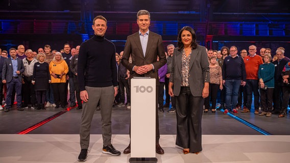 Im Vordergrund stehen Alexander Bommes, Ingo Zamperoni und Düzen Tekkal an einem Pult. Hinter ihnen stehen 100 Menschen verschiedenen Alters und Geschlechts. © NDR 