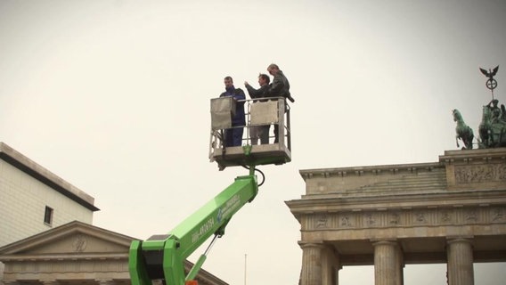 In einem Krankorb stehen drei Menschen, im Hintergrund ist das Brandenburger Tor zu sehen.  