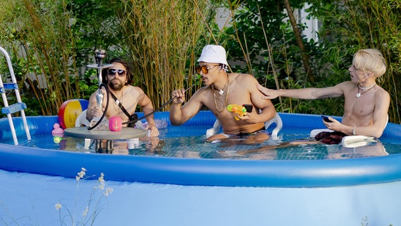 Drei junge Männer mit Sonnenbrillen baden in einem Pool - Szene aus "Player of Ibiza" © NDR/Hannah Aders Foto: Hanna Aders