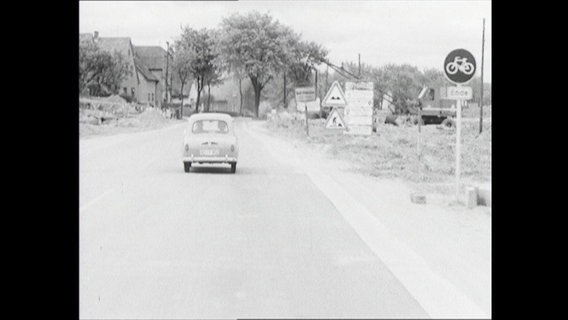 Ein Auto fährt auf einer Landstraße in ein Dorf (1964)  