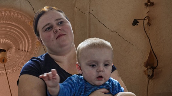 Thumbnail: Eine Frau hat ein kleines Kind in ihren Armen, im Hintergrund eine Wand mit Rissen © Screenshot 