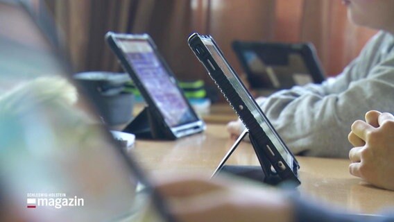 Auf einem Schultisch stehen mehrere iPadsm dazwischen die aufgestützten Arme von Schulkindern. © Screenshot 