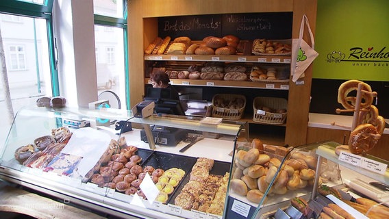 An der Verkaufstheke einer Bäckerei steht eine Verkäuferin, hinter und vor ihr liegen in der Auslage frische Backwaren. © Screenshot 