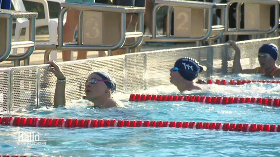 Mehrere Schwimmende halten sich in einem Sportschwimmbecken am Beckenrand unter den Startblöcken fest. © Screenshot 