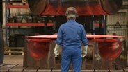 Ein Arbeiter in einem Blaumann steht vor einem größeren Rohr, das in einer Fabrik geformt wird. © Screenshot 