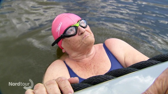 Eine ältere Dame mit rosa Schwimmkappe und Schwimmbrille hält sich im Wasser an einem Bootsrand fest. © Screenshot 