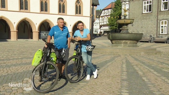 Zwei Fahrradfahrer lehnen sich an ihre auf einem Markplatz in einer Altstadt geparkten Räder. © Screenshot 