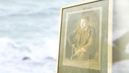 Ein altes Foto in einem Bilderrahmen auf dem ein Mann abgebildet ist, im Hintergrund das Meer. © Screenshot 