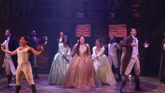 In einer Musicalszene aus "Hamilton" tanzen und singen mehrere Menschen auf einer Bühne. © Screenshot 