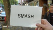 Eine Hand hält einen Zettel mit der Aufschrift "Smash" in die Kamera. © Screenshot 