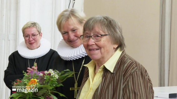 Zwei Frauen in kirchlichen Gewändern überreichen einer älteren Frau einen Blumenstrauß. © Screenshot 