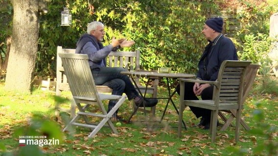Zwei Männer sitzen in einem Garten an einem Tisch und unterhalten sich. © Screenshot 