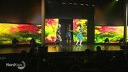Auf einer Bühne tanzen drei Darstellende in bunten Kostümen. © Screenshot 