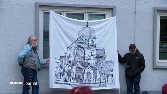 Zwei Männer halten ein Transparent, auf dem eine Synagoge gezeichnet ist. © Screenshot 