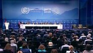 Der Konferenzsaal der Weltklimakonferenz mit zahlreichen Personen auf und vor der Bühne des Saals © Screenshot 
