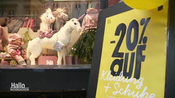 Ein gelbes Schild steht vor einem Schaufenster eines Kinderbekleidungsladens und kündigt Prozente an. © Screenshot 