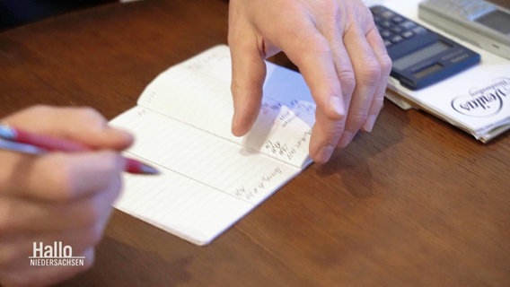 Nahaufnahme von zwei Händen, die etwas in ein Kassenbuch reinschreiben. Daneben liegt ein Taschenrechner. © Screenshot 