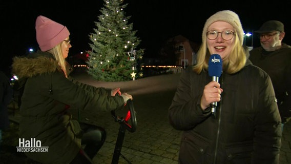Reporterin Marie Schiller neben einer Frau auf einem Sportgerät, im Hintergrund ein beleuchteter großer Tannenbaum. © Screenshot 