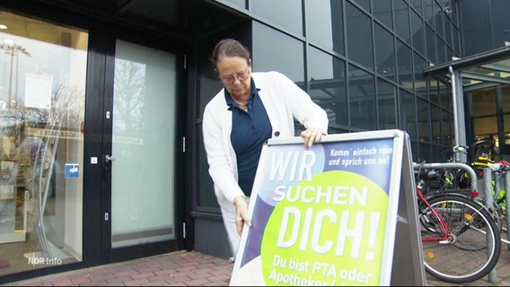 Eine Apothekerin stellt ein Schild vor der Eingangstür auf, auf dem steht: Wir suchen dich! © Screenshot 