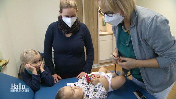 Eine Ärztin untersucht ein Baby, die Mutter und die Schwester stehen daneben. © Screenshot 