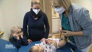 Eine Ärztin untersucht ein Baby, die Mutter und die Schwester stehen daneben. © Screenshot 