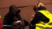 Eine Ehrenamtliche vom Hamburger Kältebus kniet neben einem Mann der obdachlos ist. Er hat die Kapuze seiner Jacke tief ins Gesicht gezogen und hält ein warmes Getränk in den Händen. © Screenshot 