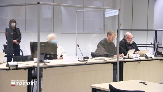 Im Gerichtssaal: Die Angelagte (unkenntlich gemacht) und ihre Verteidiger, im Hintergrund steht eine Polizeibeamtin. © Screenshot 