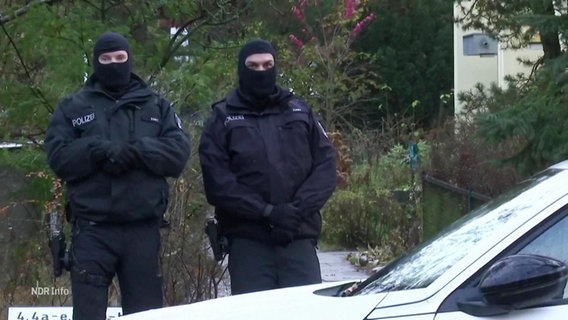 Zwei vermumte Polizisten stehen vor einem Haus. © Screenshot 