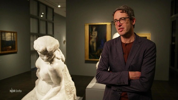 Ein Mann in Anzug und Brille steht neben der Skulptur einer nackten Frau. © Screenshot 