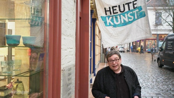 Ein NDR-Reporter steht vor einem Geschäft, an dem eine Fahne mit der Aufschrift "Heute Kunst" hängt. © Screenshot 