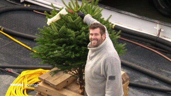 Eine Person aus der Schifffahrt mit einem Weihnachtsbaum in der Hand, die grinst. © Screenshot 