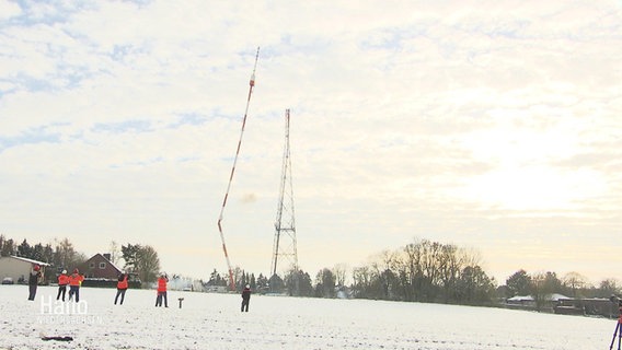 Ein umknickender Turm auf einem schneebedeckten Feld, daneben ein weiterer Sendemast und Zuschauende. © Screenshot 