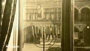 Eine historische Aufnahme zeigt einen jüdischen Chanukka-Leuchter vor einem Haus an dem eine NS-Flagge hängt. © Screenshot 