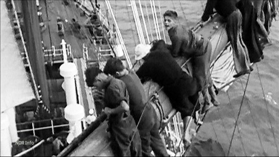 Eine Personen hängen am Mast eines Schiffes. Bild in Schwarz/Weiß © Screenshot 