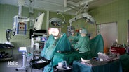 Krankenhauspersonal steht eingekleidet in hyginische Schutzausrüstung an einem OP-Tisch im Operationssaal. © Screenshot 