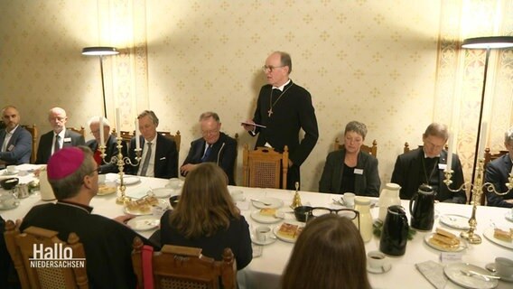 Mehrere Menschen sitzen an einer langen Tafel. In der Mitte steht ein Bischof und spricht. © Screenshot 