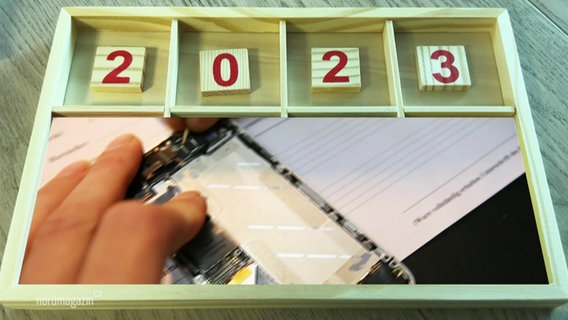 Ein Holzkasten, in dem die Zahlen "2023" liegen. Im unteren Segment ist ein Bild einer Hand mit einem kaputten Handy zu sehen. © Screenshot 