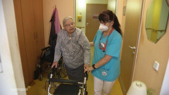 Eine Altenpflegerin führt eine Seniorin am Arm durch ihr Zimmer. © Screenshot 