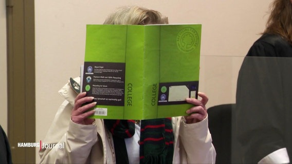Eine Angeklagte vor Gericht hält sich ein dickeres Buch mit grünem Buchrücken vors Gesicht. © Screenshot 